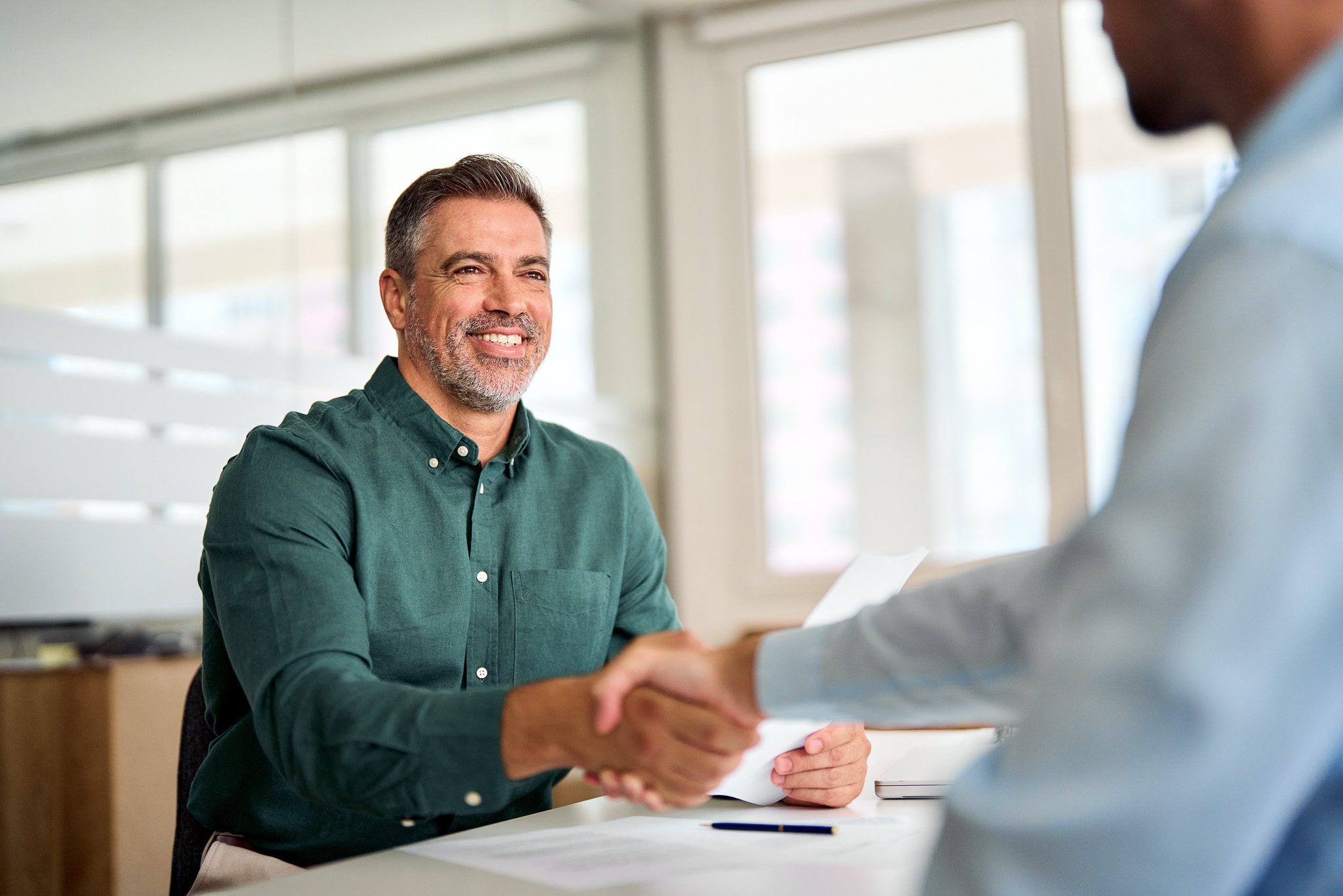 Smiling,Middle,Aged,Business,Man,Handshaking,Partner,Making,Partnership,Collaboration A man in a green shirt smiles while shaking hands with another person in an office setting.