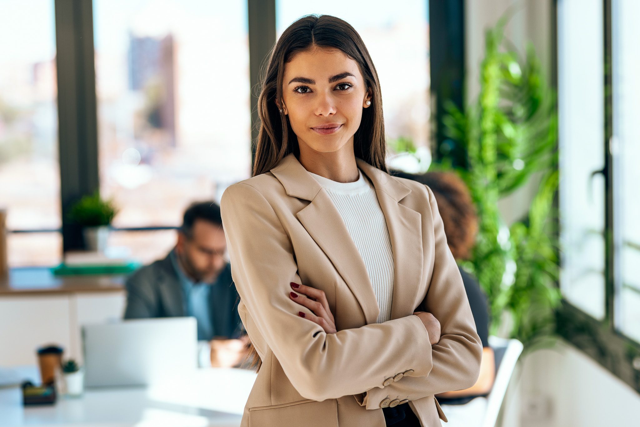 Portrait,Of,Beautiful,Young,Business,Woman,Standing,While,Smiling,Looking A woman in a beige blazer stands confidently with arms crossed in a bright office. Two people work on laptops in the background.